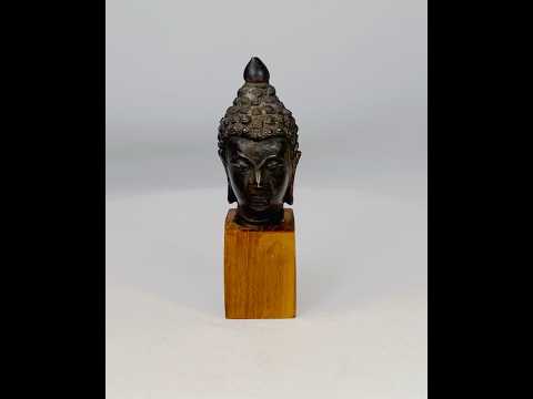 Antique Southeast Asian cast bronze Buddha head, likely from Thailand or Burma, 18th–19th Century CE. Features serene facial expression, closed eyes, gentle smile, ushnisha symbolizing wisdom, and intricately modeled hair curls. Dark natural patina with wooden base, reflecting Theravāda Buddhist devotional artistry and meditation iconography.