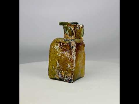 Early Imperial Roman mold-blown glass bottle from the 1st Century CE, likely Rhineland. Square translucent body with cylindrical neck, flaring folded rim, and wide ribbed “celery handle.” Base bears four-leaf maker’s mark. Developed iridescent patina reflects age and Roman glassmaking innovation, used for oils, perfumes, or wine.