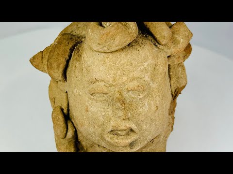 Ancient Pre-Columbian terracotta head from Mesoamerica, featuring stylized facial features with closed eyes, parted lips, and remnants of elaborate headdress elements, hand-molded from clay, dating between 1000–1500 CE, likely used as a ritual offering or funerary effigy.