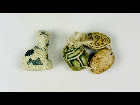 Ancient Egyptian faience cat amulet with traces of blue-green glaze, shown alongside three scarabs in varied materials, on a white background.