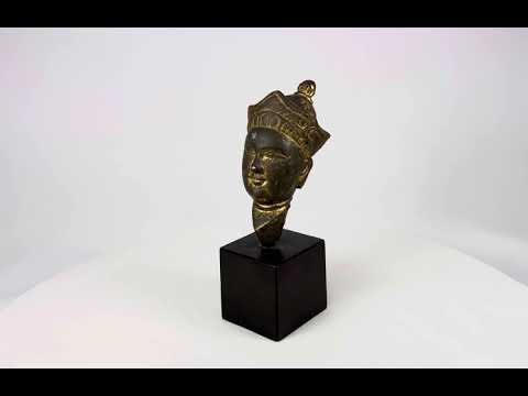 Antique Tibetan gilded bronze Buddha head with ornate crown, serene facial features, and traces of gilding, mounted on a black display base, photographed on a white background.