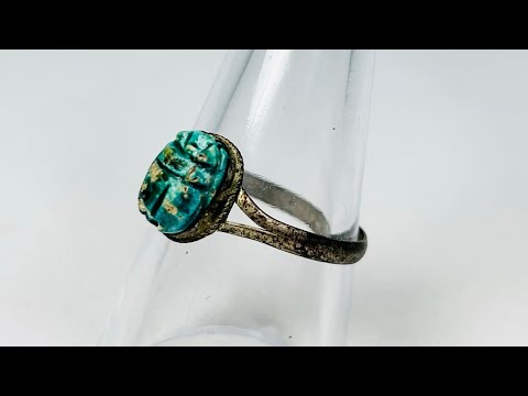 Ancient Egyptian scarab ring with turquoise-green faience scarab set in bronze band, displayed on a clear stand against a white background.
