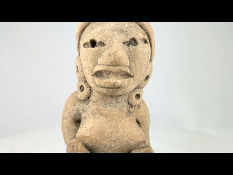 Ancient West Mexican terracotta female figurine, seated with earspools and headdress, hand-modeled with expressive facial features, mounted on a black display base, photographed on a white background.