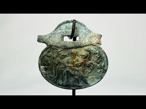 Ancient Roman bronze pendant, 1st–4th century CE, engraved with three standing figures including a helmeted figure, green patina, mounted on a custom stand.