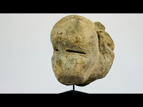 Ancient Mesoamerican stone mask fragment with minimalist facial features and narrow eye slits, carved from a single block of stone and mounted on a black wooden display stand, dating to 500 BCE–500 CE.