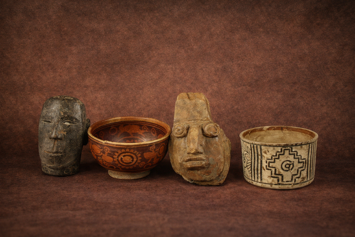 Professional horizontal gallery photograph featuring four ancient artifacts — Polynesian volcanic stone head, Pre-Columbian red-slip pottery bowl, Valdivia anthropomorphic spouted jar, and Indus Valley painted pottery vessel — displayed against a warm museum-style brown backdrop.