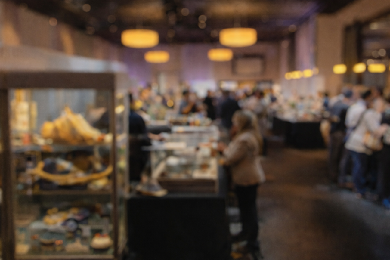 Relic & Rarity attending an antique and artifact exhibition with softly blurred antique and artifacts, glass display cases and collectors viewing historical objects under warm gallery lighting.