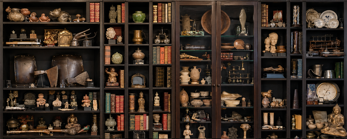 Wide banner of a dark wood display cabinet filled with curated historical artifacts, including sculptures, pottery, books, and metal objects arranged in a museum-style collection.