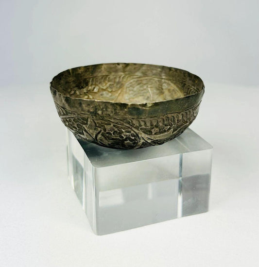 Angled view of Seljuk Persian silver unguent bowl from 12th–13th century CE, featuring intricate engraved floral and geometric motifs with aged silver patina, displayed on a clear acrylic base under studio lighting.